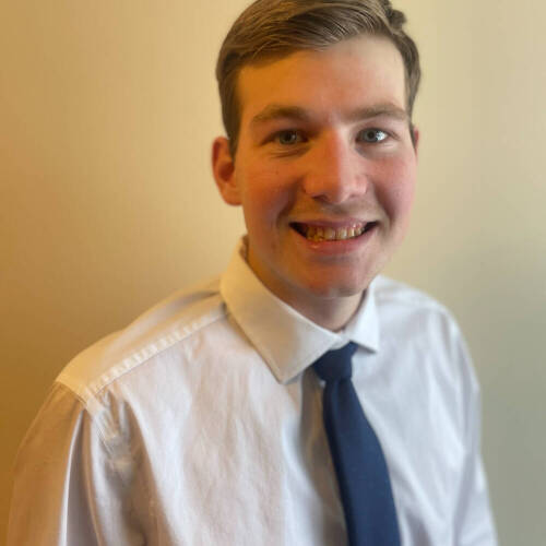 Colin Nagley in a white collared shirt with blue tie on a yellow backdrop.